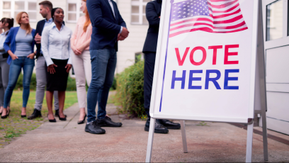 A vote here sandwich board sign with people standing in line waiting to vote.