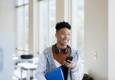 Male college student in lecture room, smiling and using a smartphone.