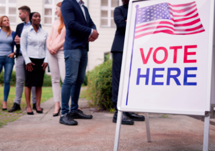 A vote here sandwich board sign with people standing in line waiting to vote.