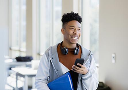 Male college student in lecture room, smiling and using a smartphone.