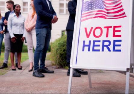 A vote here sandwich board sign with people standing in line waiting to vote.
