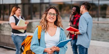 Smiling female student wearing glasses and carrying a yellow backpack and blue folder.