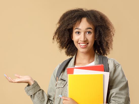 Smiling female student with backpack and notebooks points to empty space and looks at camera, yellow background