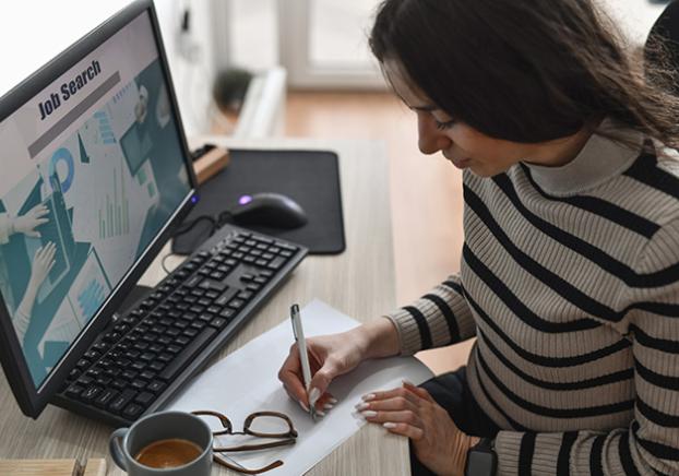 Woman sitting at a computer searching for jobs and taking notes