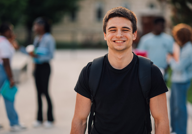 Male student in a black t-shirt standing at campus and smiling at camera.
